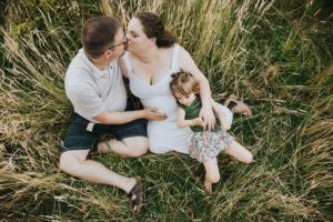 Pregnant mom, dad/partner, and oldest daughter snuggle and tickles in the grass during their maternity session in Hillsboro, Oregon