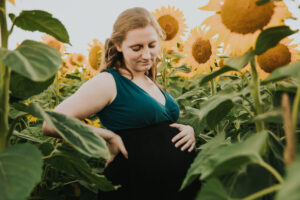 Redheaded pregnant mom holds her bump in the front and looks down her chest, framed by sunflowers in Hillsboro Oregon