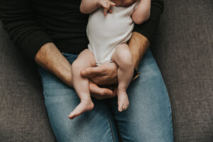 Close up on dad's arms holding baby in lap on the couch during lifestyle newborn session by Portland Newborn Photographer.