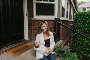 Mom's holds baby in lap and smiles sitting on their apartment doorstep Baby held outdoors in mom's lap during lifestyle newborn session by Portland Newborn Photographer.