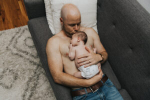 Shirtless dad on the couch holds baby on his chest and kisses her head during lifestyle newborn session by Portland Newborn Photographer.