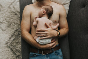 Close up on baby sleeping on shirtless dad chest on the couch Shirtless dad on the couch holds baby on his chest and kisses her head during lifestyle newborn session by Portland Newborn Photographer.
