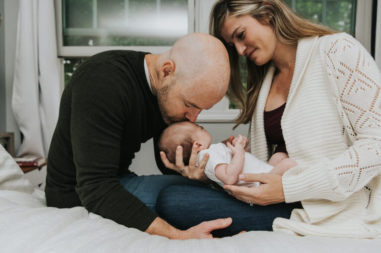 Mom looks on as dad bends over to kiss baby on forehead on their bed at a lifestylew newborn session by portland newborn photographer.