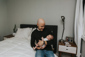 Dad smiles holding an open eyed baby on a bed during lifestyle newborn session by Portland Newborn Photographer.