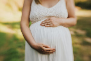 Close up on a slightly out-of-focus pregnancy bump in a white dress with both hands holding during a maternity session in Yamhill, Oregon
