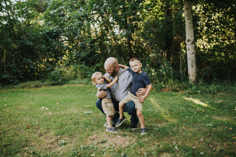 Dad crouches down in a nature setting and has 2 boys sitting on his lap during a maternity session in Yamhill, Oregon