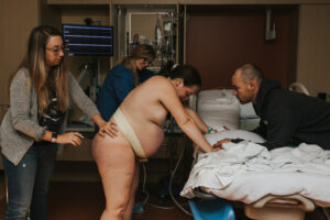 Birthing person bend overs a hospital bed at Kaiser Westside and holds her partner's hands while a doula providers physical support.