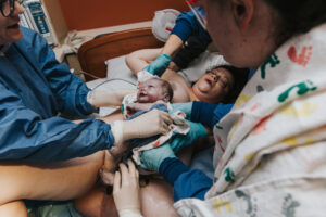 Birthing mom is laying on the hospital bed and newly birthed baby is being wiped down by several hands as he is being put onto her chest at Kaiser Westside Hospital in Hillsboro Oregon
