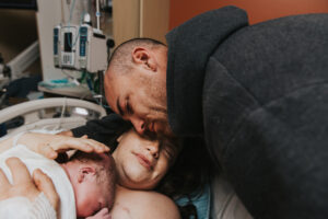 A new dad with tears in his eyes leans over and leans his head against the birthing mom's who has her arms wrapped around their baby