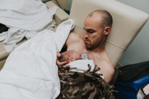 New dad looks down, marvels at his new baby during skin to skin, and cups his head gentle with a few blankets on top of them