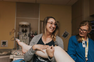 Labor doula Kendelle looks to the side and smiles big while holding a side lying birthing mom's top leg up high while nurses attends