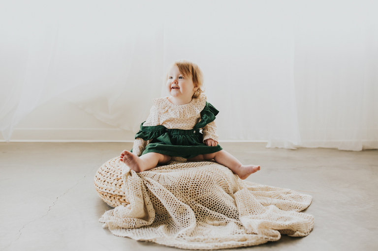 Nora the redheaded tot in a green jumper smiles on top of a stool with a lace blanket during her first birthday portraits in Portland Oregon