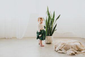 Tot Nora with a green jumper falling off her body, stands next to a large potted plant in a studio for her first birthday photos in Portland Oregon