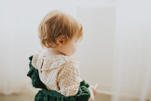 Side profile on a redheaded tot with a green jumper hanging off of her shoulder in front of a white gauze curtain in a Portland Oregon studio
