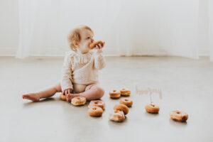 A redheaded tot sits on the floor with a donut in her mouth and donuts on the floor around her during her Portland Oregon donut smash.