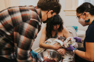 Mom sits on toilet holding baby while 2 Unfurling Birth midwives provide care post home birth in Portland Oregon