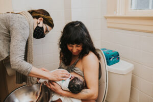 Mom sits on toilet holding baby and looking down while an Unfurling Birth midwife provides care post home birth in Portland Oregon